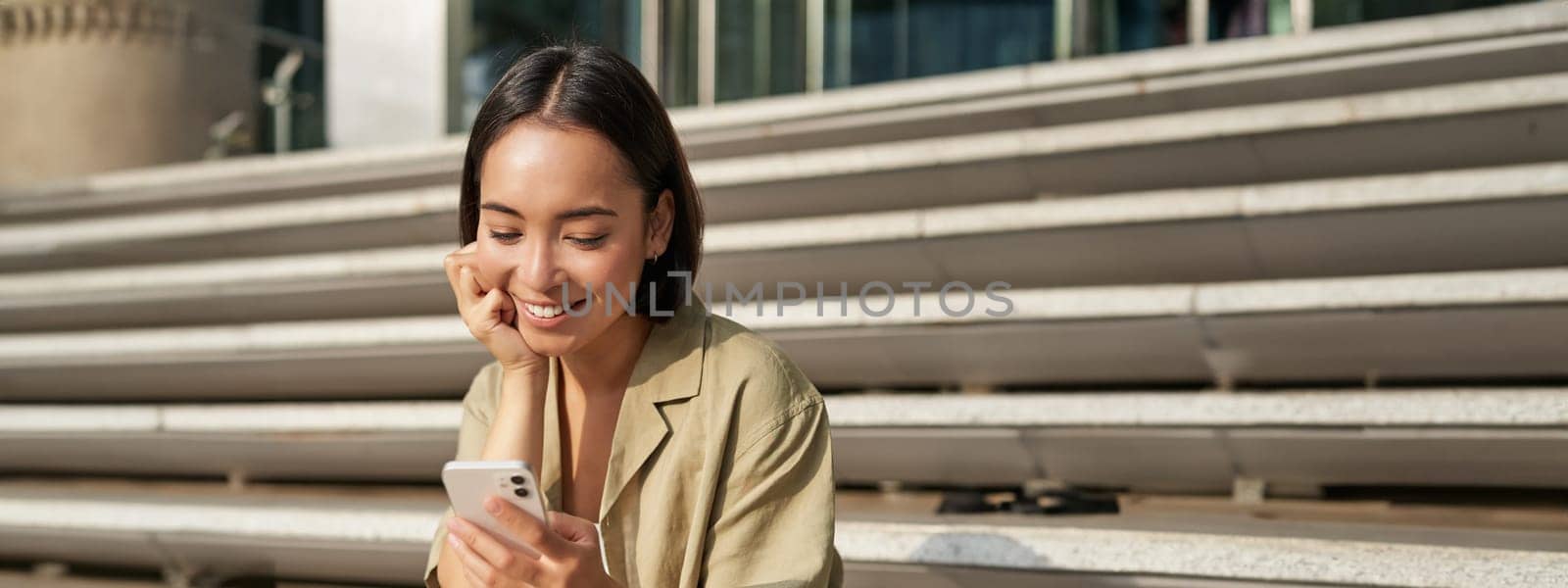 Vertical shot of asian woman, student sits on stairs in city, looking at mobile phone screen and smiling, using smartphone app.