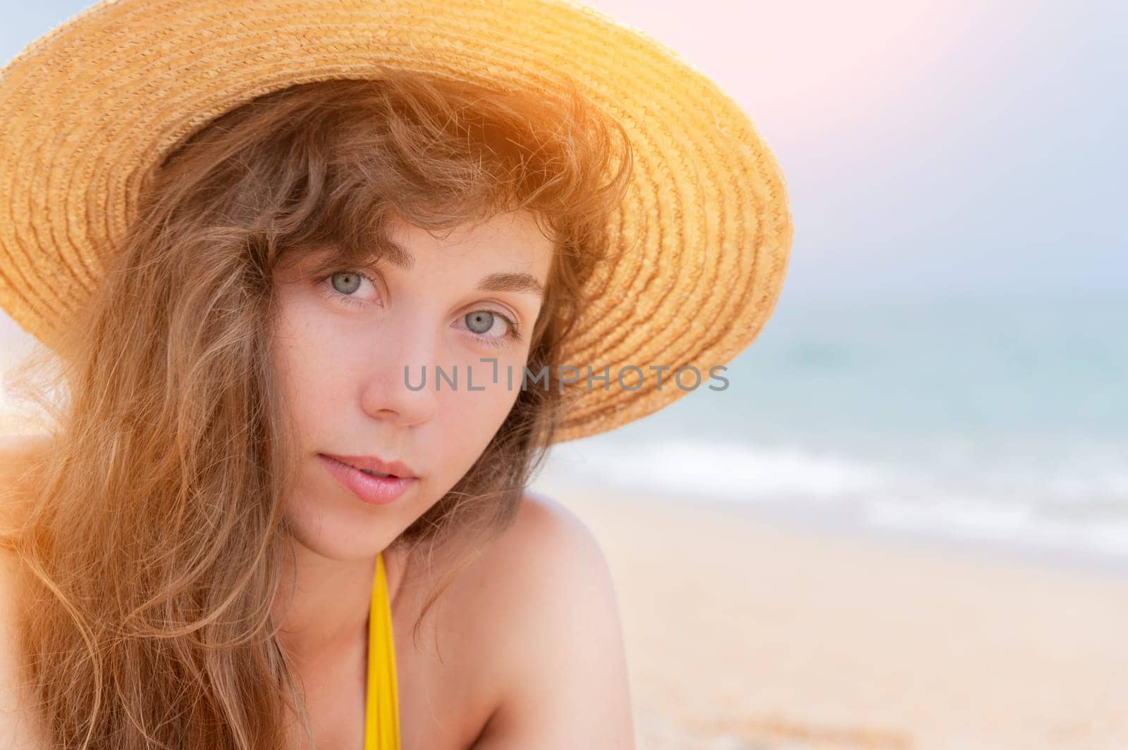 Portrait of a beautiful smiling young woman in a straw hat on the beach against the background of the ocean. The girl looks at the camera on the seashore. Carefree tanned woman lying on the sand.