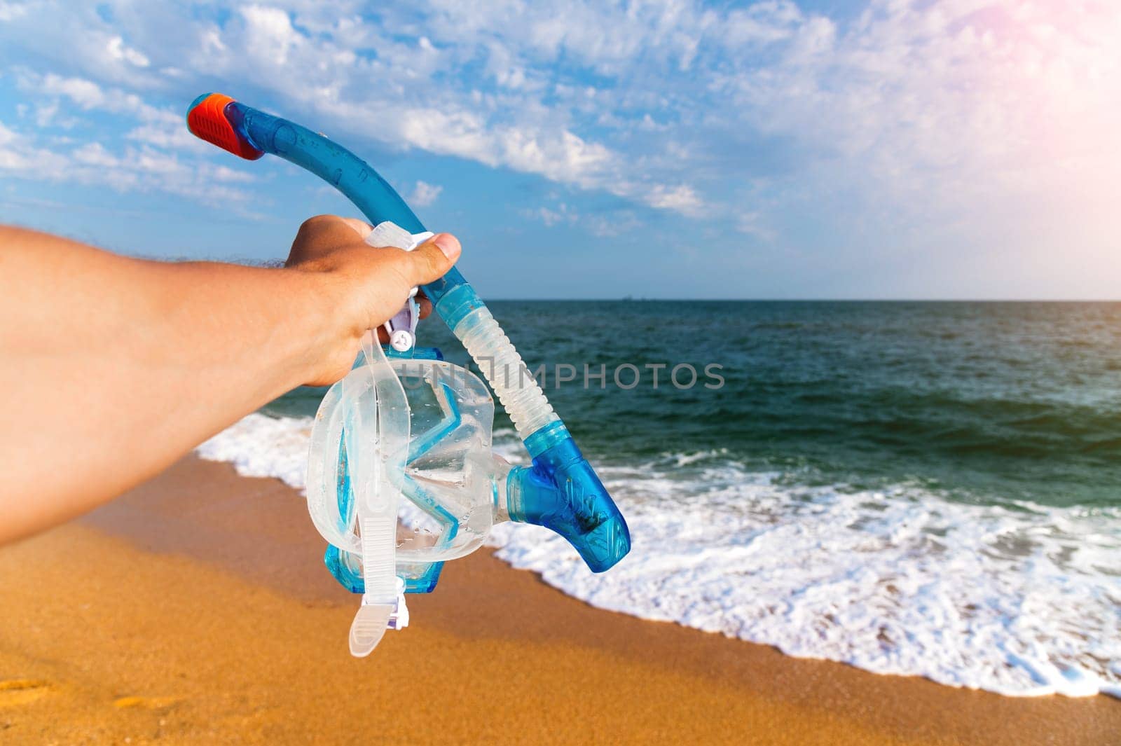 A man holds a snorkeling mask in his hand against a background of a blue sky with clouds and a turquoise ocean. Diving mask in a man's hand. Photo of mask and snorkel for swimming in the sea