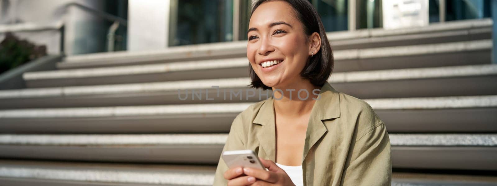 Beautiful asian girl sits on stairs with smartphone. Young korean woman resting outdoors, using mobile phone application.