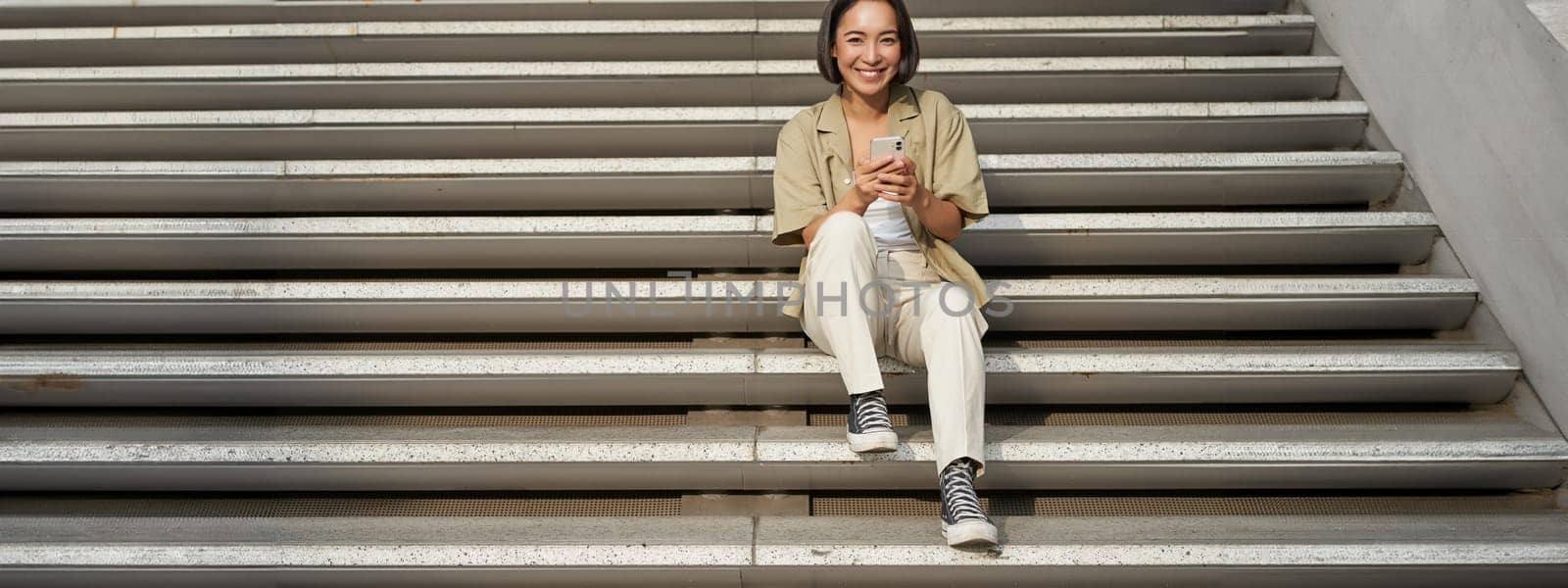 Portrait of smiling asian girl sits on stairs with her smartphone, browsing internet on mobile phone, resting outdoors in city centre.