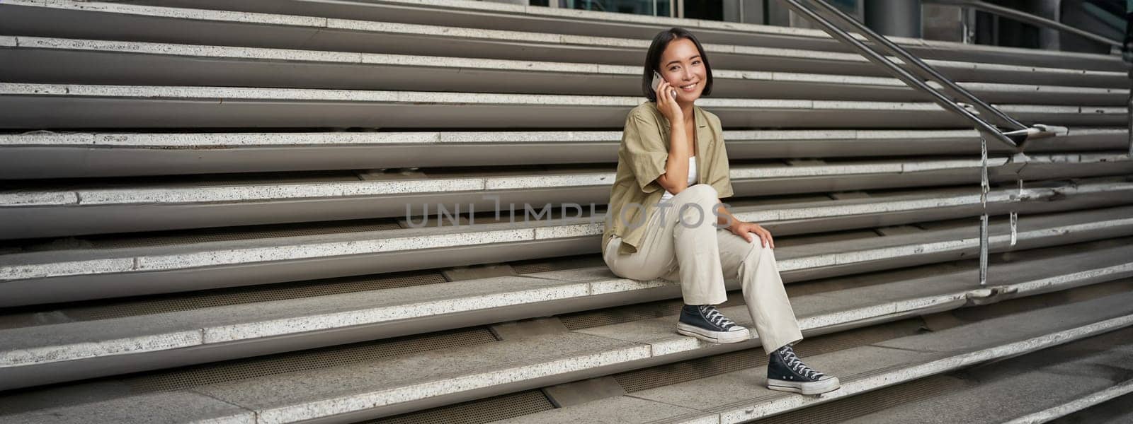 Smiling asian girl sits on stairs of building and talks on mobile phone, relaxing during telephone conversation.