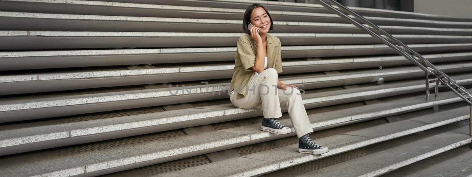 Portrait of asian student girl on stairs talks on mobile phone, smiles at camera, sits outside building.