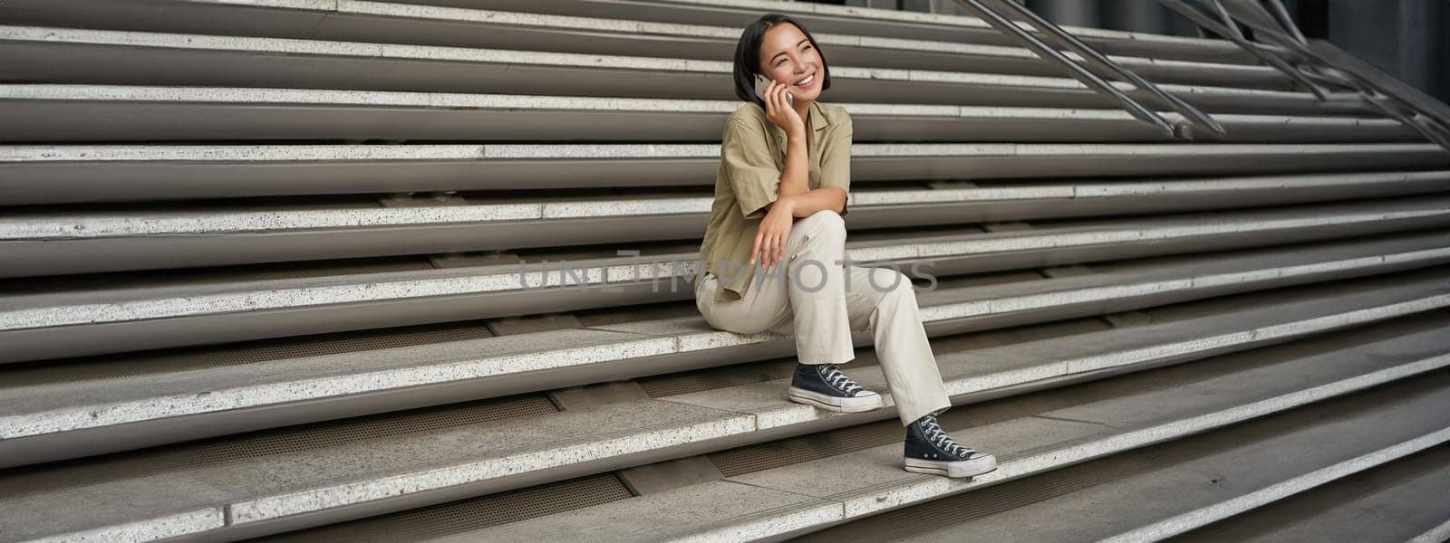 Portrait of asian student girl on stairs talks on mobile phone, smiles at camera, sits outside building.