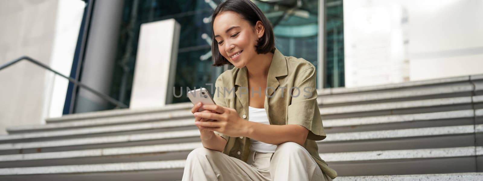 Technology and communication. Young smiling girl, asian woman sits with smartphone, reads message with big smile.