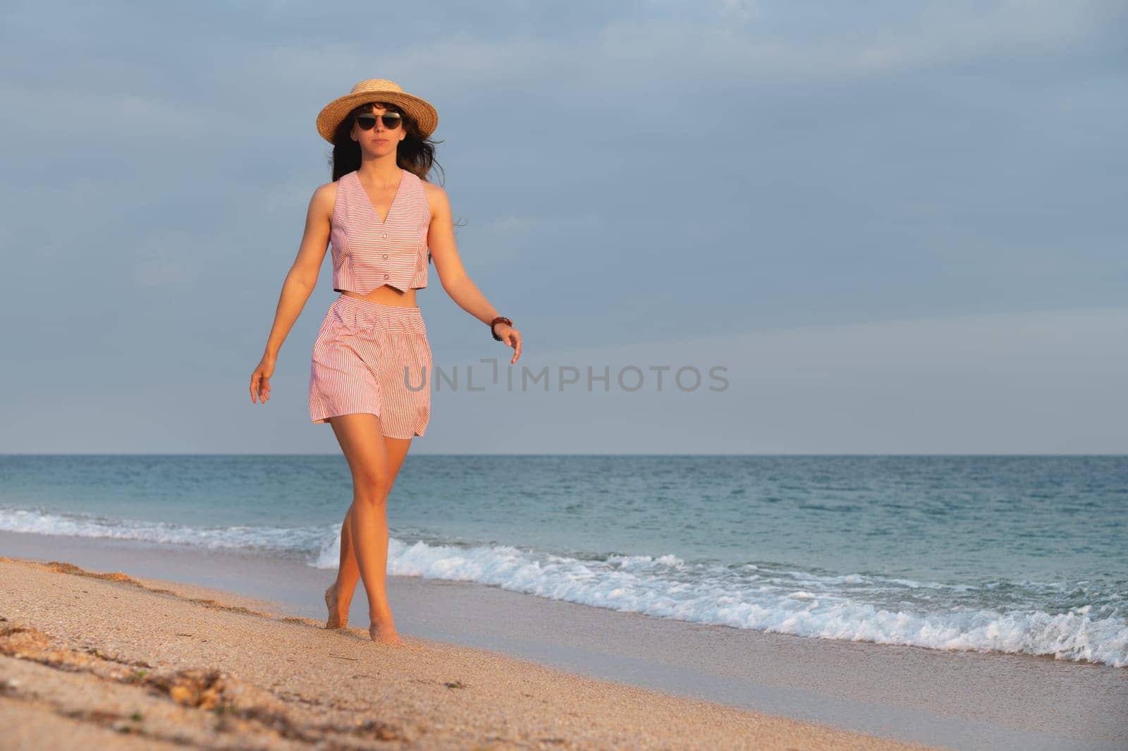 elegant woman in a suit and a straw hat on the ocean while walking at sunset. Beautiful young woman in glasses and a hat walks along the beach.