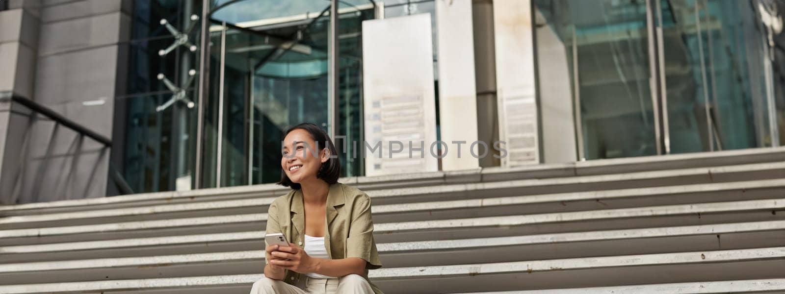 Vertical shot of happy smiling asian girl sits on stairs in city centre, holds mobile phone and looks aside with pleased face. Young woman using her smartphone outdoors.