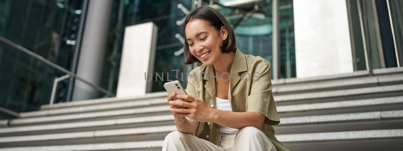 Technology and communication. Young smiling girl, asian woman sits with smartphone, reads message with big smile by Benzoix