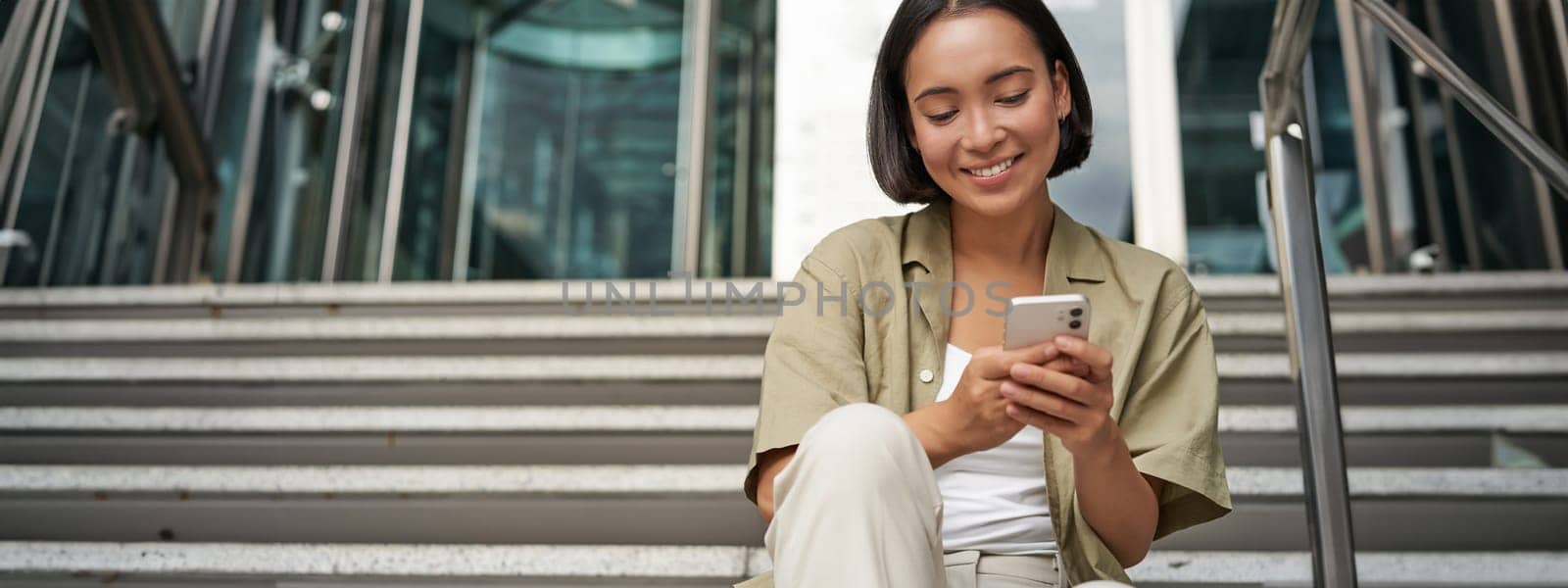 Smiling asian girl sits on stairs near building entrance, using mobile phone app. Happy young woman rests with smartphone in her hands outdoors by Benzoix