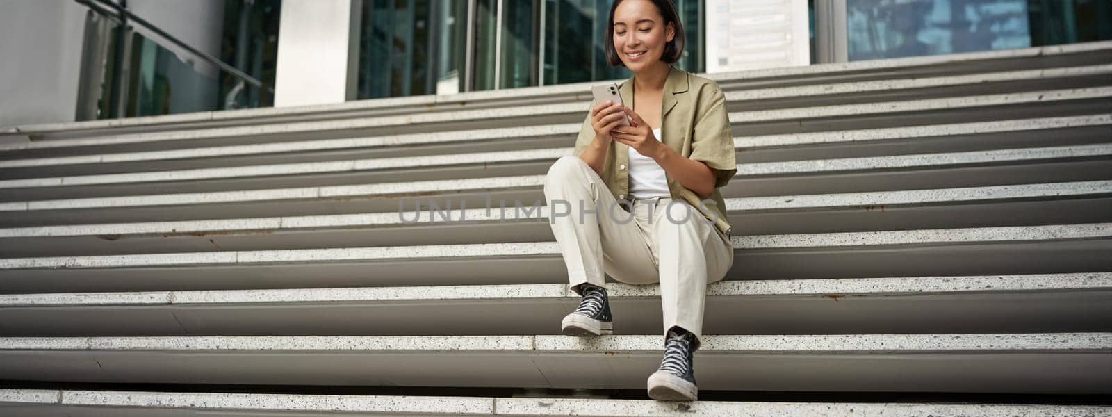 Cellular technology and people. Young happy asian girl sits with smartphone in front of building. Woman using mobile phone, smiling while looking at screen.