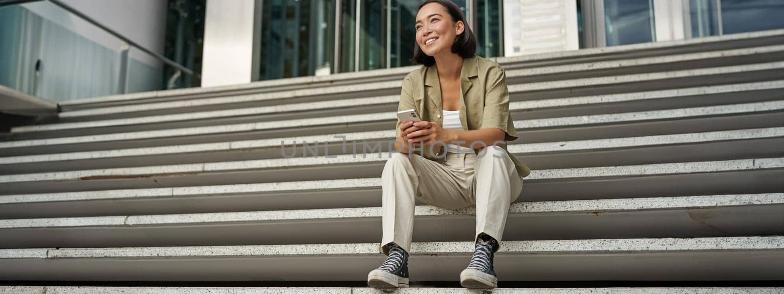 Portrait of happy smiling asian woman, sitting outdoors near building, using smartphone. Technology concept by Benzoix