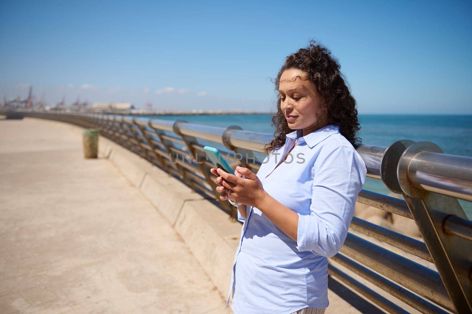 Charming multi-ethnic woman in blue casual shirt using smartphone and mobile app, checking social media content, booking, ordering, scrolling newsfeed, smiling, enjoying beautiful sunny day