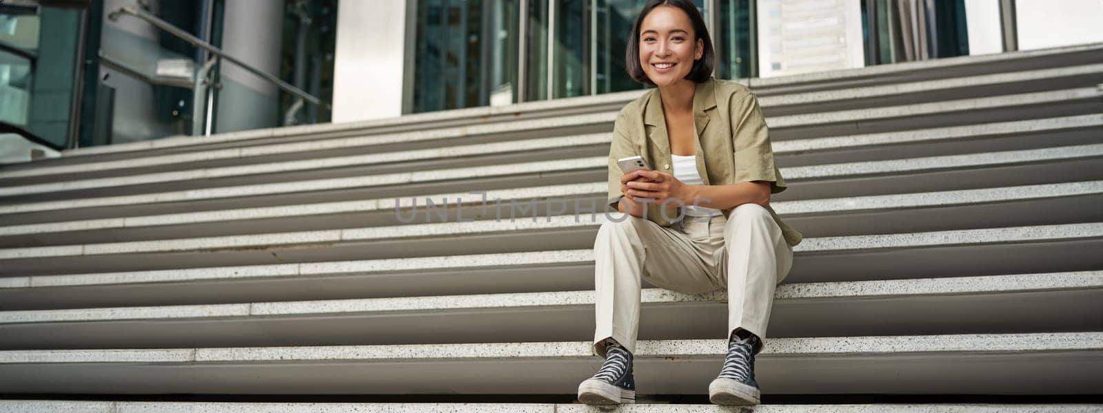 Portrait of smiling asian girl sits on stairs outdoors, sending message, using smartphone app, looking at mobile phone screen.