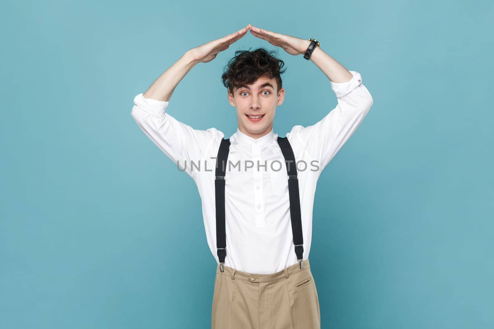 Happy to feel safe. Portrait of man wearing white shirt and suspender showing house roof symbol over head, smiling joyfully, gesture of protection. Indoor studio shot isolated on blue background.
