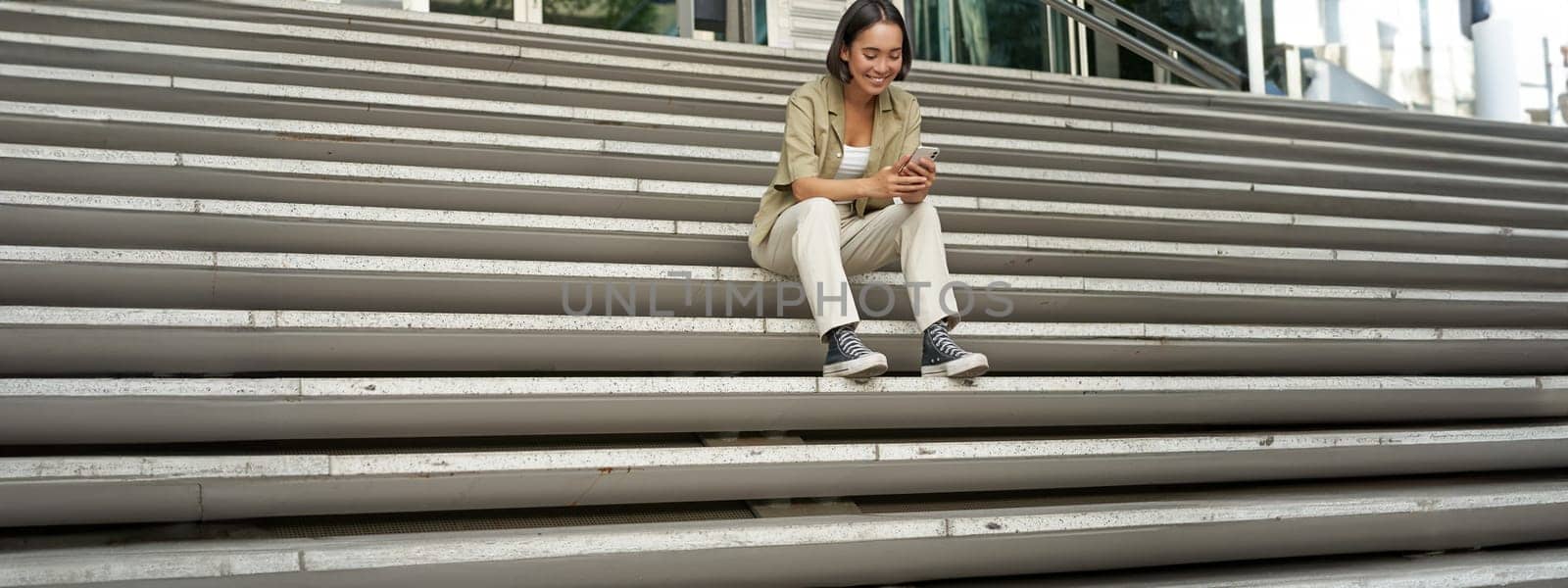 Portrait of smiling asian girl sits on stairs outdoors, sending message, using smartphone app, looking at mobile phone screen by Benzoix