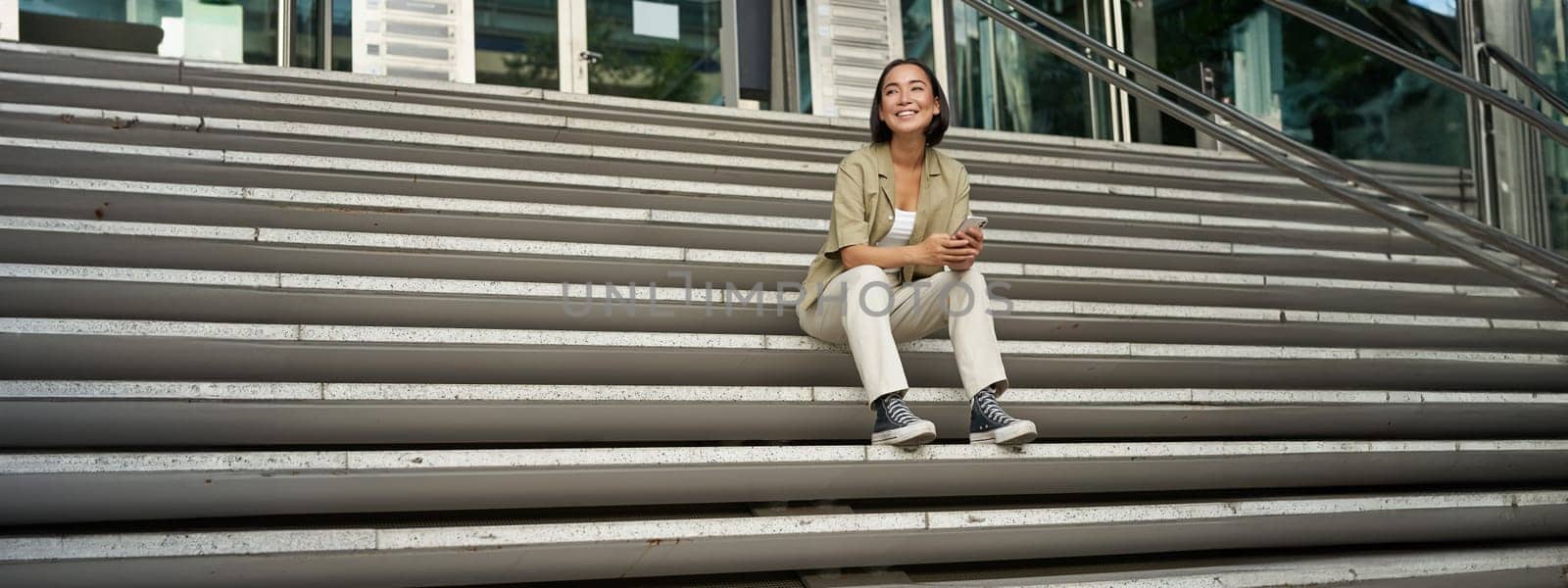 Portrait of smiling asian girl sits on stairs outdoors, sending message, using smartphone app, looking at mobile phone screen by Benzoix