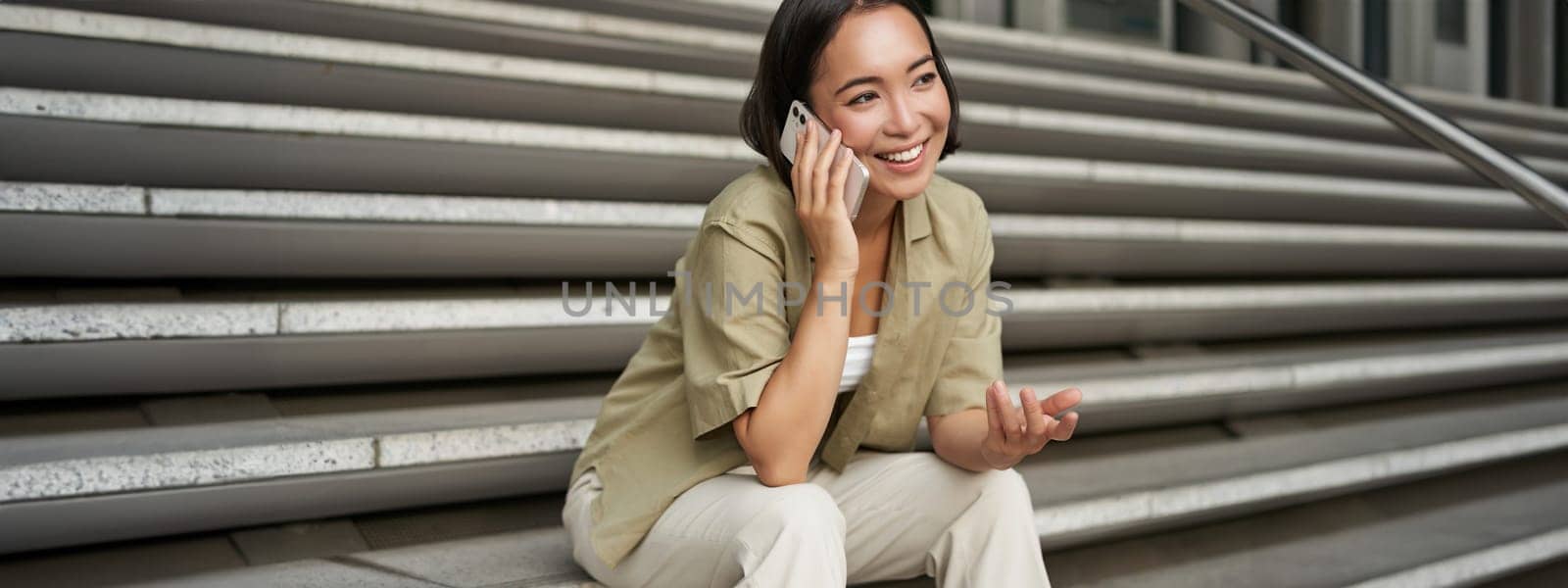 Close up portrait of smiling asian girl talks on mobile phone, sits outside on street stairs. Young woman calling friend on smartphone by Benzoix