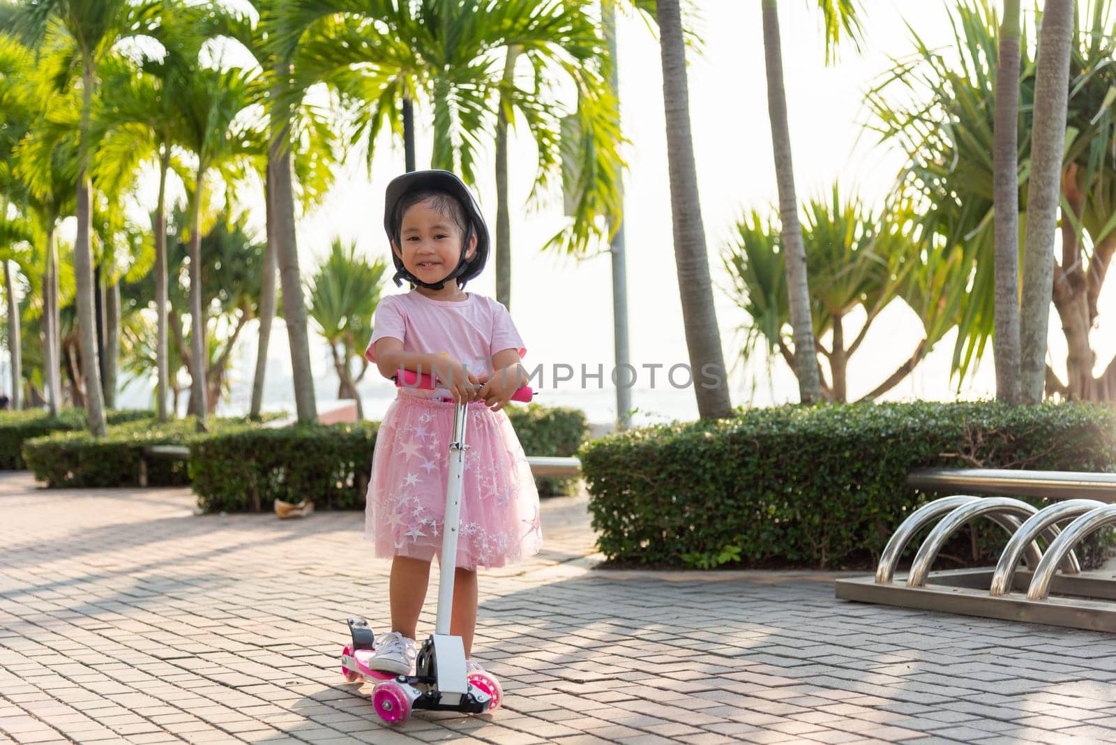 Happy Asian little kid girl wear safe helmet playing pink kick board on road in park outdoors on summer day by Sorapop