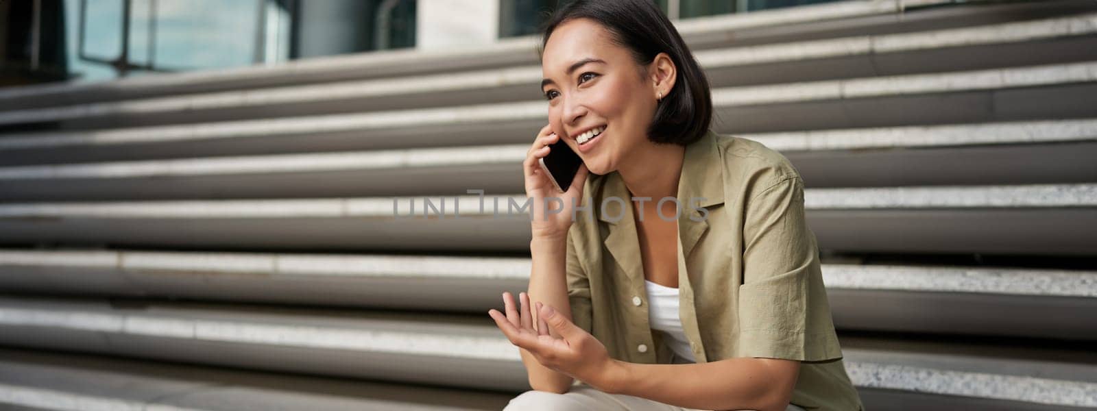 Portrait of beautiful asian girl talks on mobile phone, sits on street stairs. Woman with smartphone smiling, making a call.