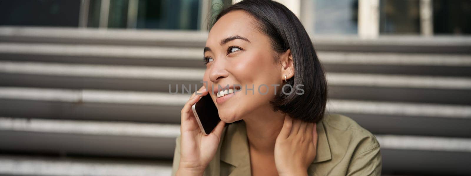 Cellular technology. Smiling asian girl talks on mobile phone, makes a call. Young woman with telephone sits on stairs.