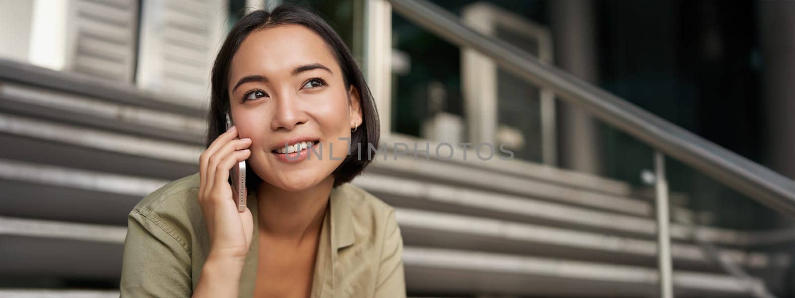 Portrait of asian girl smiles while talks on mobile phone. Young woman calling a friend, sitting on stairs by Benzoix