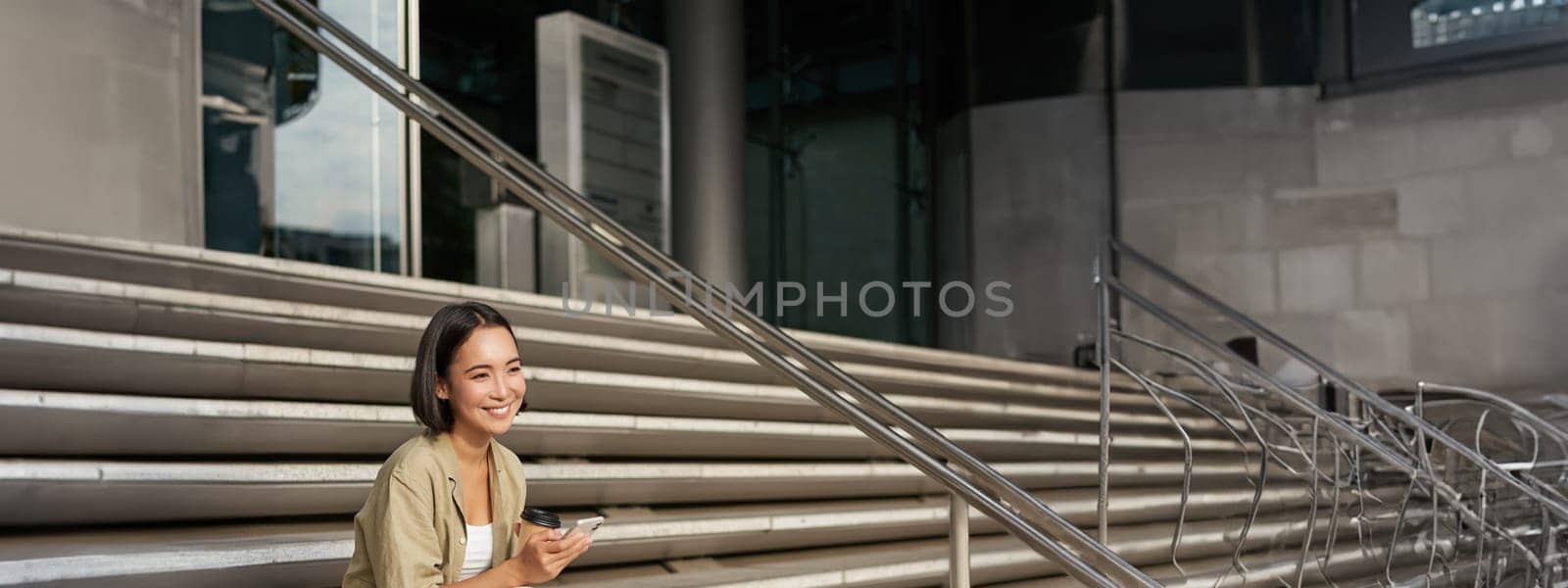 Vertical shot of young smiling woman sits on stairs in city centre wth mobile phone. Girl with telephone, browsing on smartphone, texting message by Benzoix