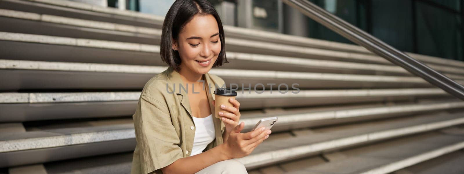 Happy urban girl drinks her takeaway coffee and scrolls feed on smartphone. Asian woman sits on stairs with tea and holding mobile phone by Benzoix