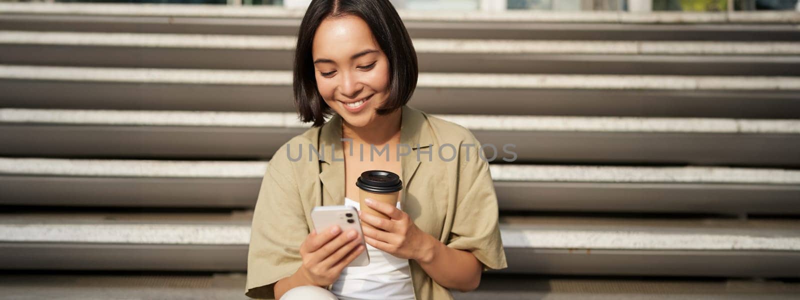Happy urban girl drinks her takeaway coffee and scrolls feed on smartphone. Asian woman sits on stairs with tea and holding mobile phone by Benzoix
