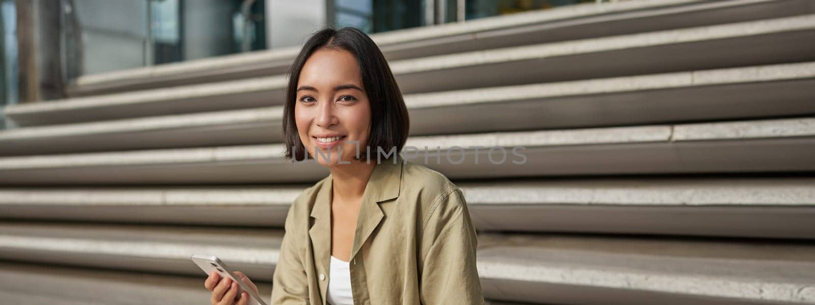 Vertical shot of asian girl drinking her coffee and using mobile phone. Young woman with smartphone and takeaway sits on stairs.