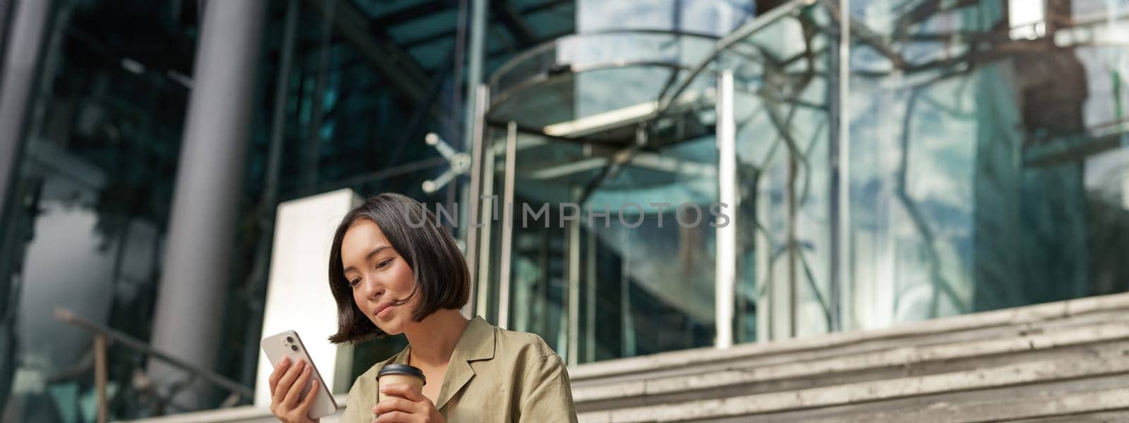 Beautiful smiling asian girl, drinking coffee, using mobile phone and sits on stairs outside. Young woman video chat outdoors by Benzoix