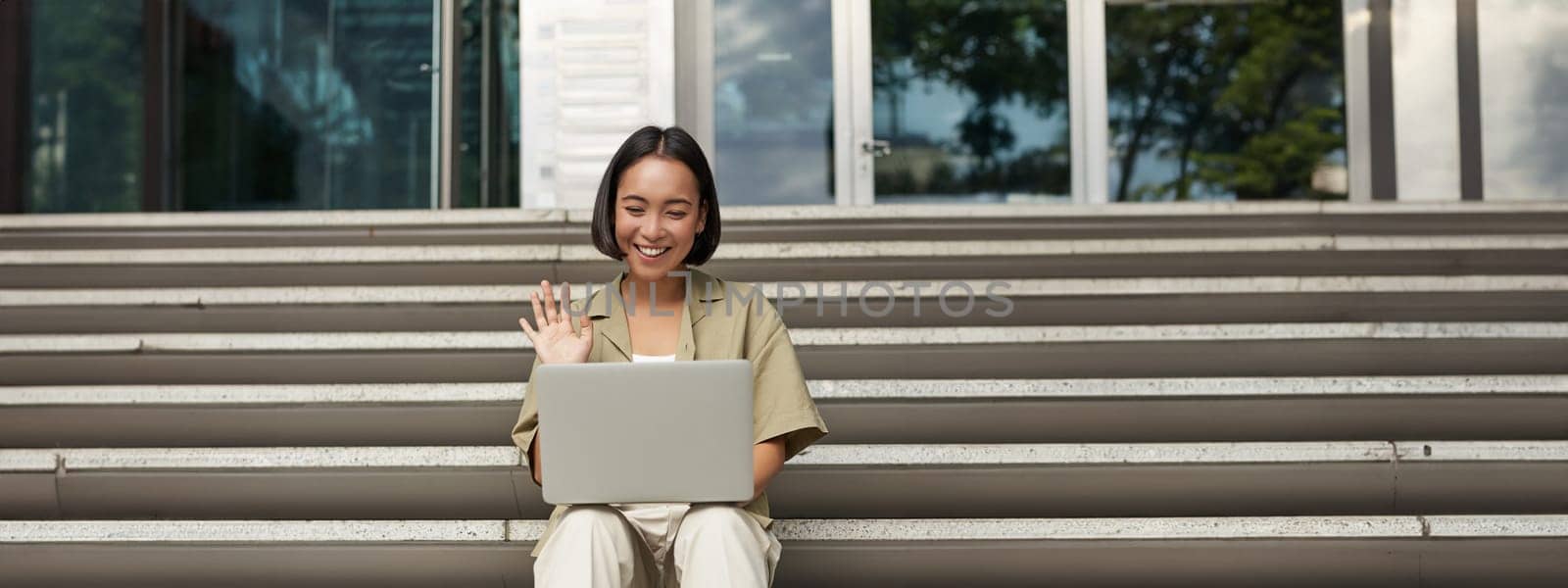 Portrait of young asian girl, student talks at laptop, video chat, speaking during online meeting, sitting outdoors on stairs.