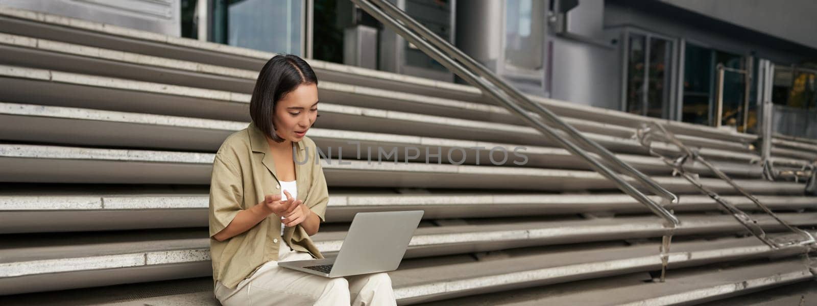 Vertical shot of asian girl student attend online meeting, talking at laptop, video chat, sitting on stairs.