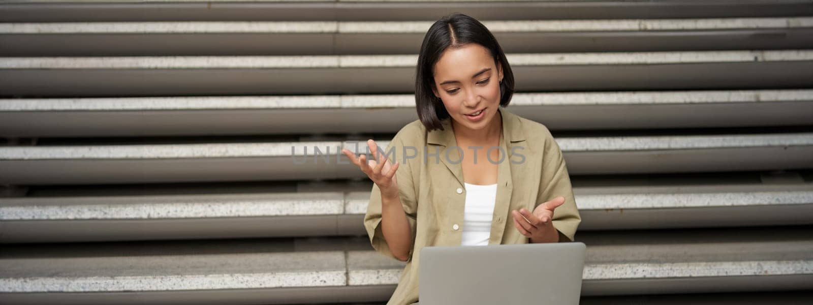 Portrait of young asian girl, student talks at laptop, video chat, speaking during online meeting, sitting outdoors on stairs.