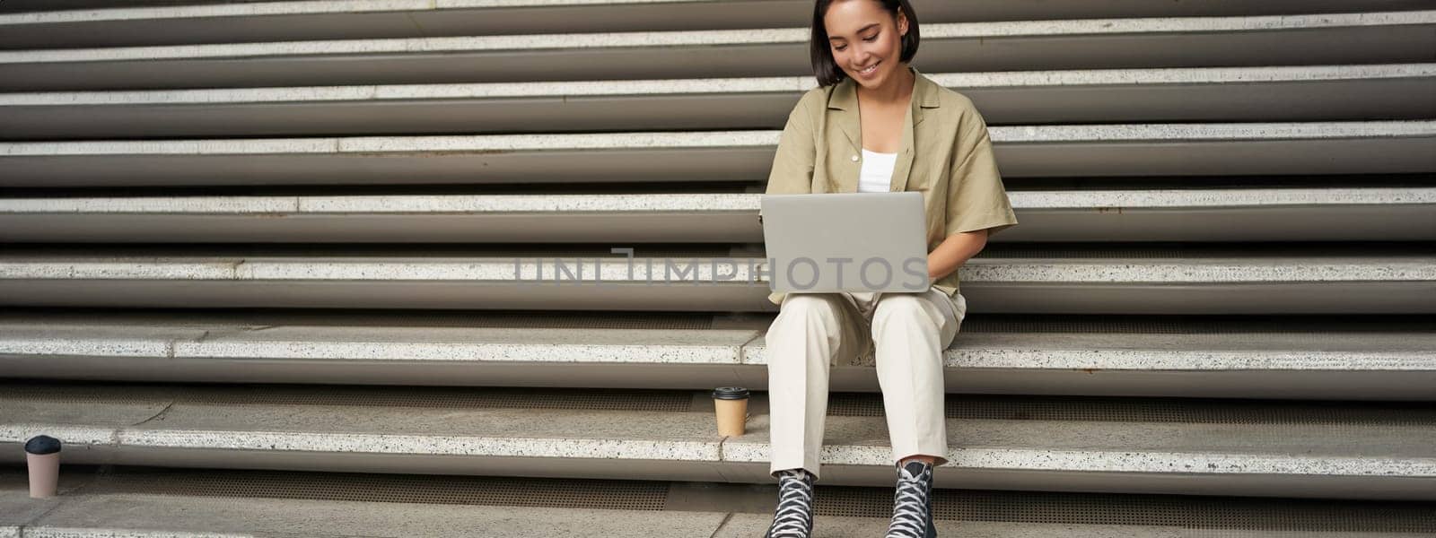 Portrait of young asian woman using her laptop, sitting outdoors on stairs. Happy smiling girl with personal computer.