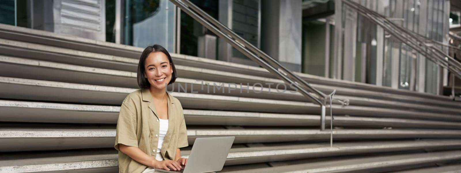 Vertical shot of asian girl sits with laptop, drinks coffee on university stairs. Young woman, student does her homework outdoors.