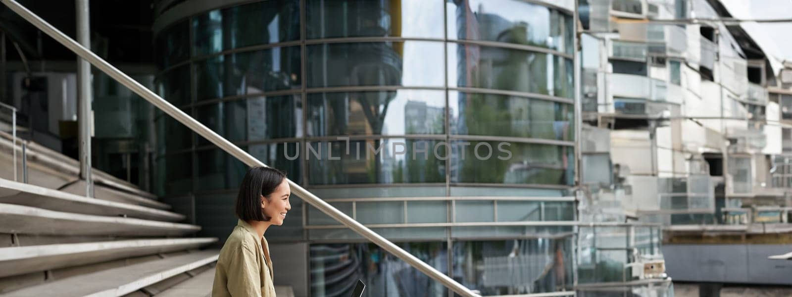 Profile portrait of young asian woman with laptop, girl student sits on stairs outside building and types on computer, drinks takeaway coffee.