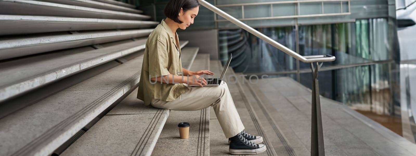 Profile portrait of young asian woman with laptop, girl student sits on stairs outside building and types on computer, drinks takeaway coffee.