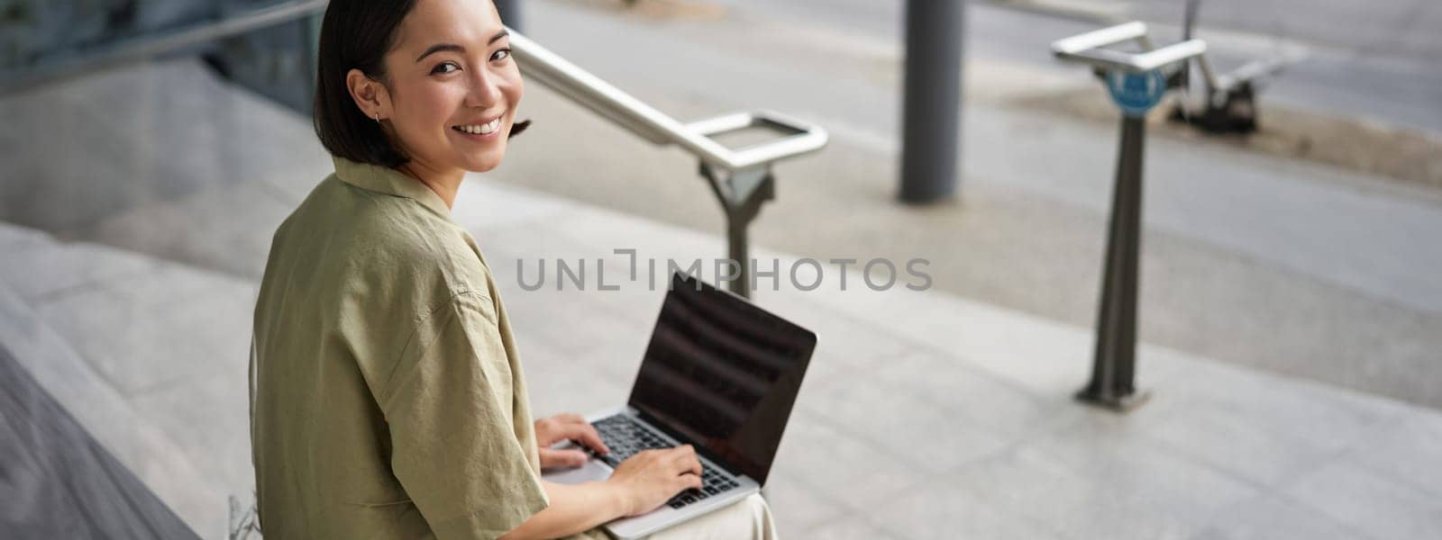 Young smiling woman, asian digital nomad using computer outdoors. Student works on project with laptop, sits on stairs.
