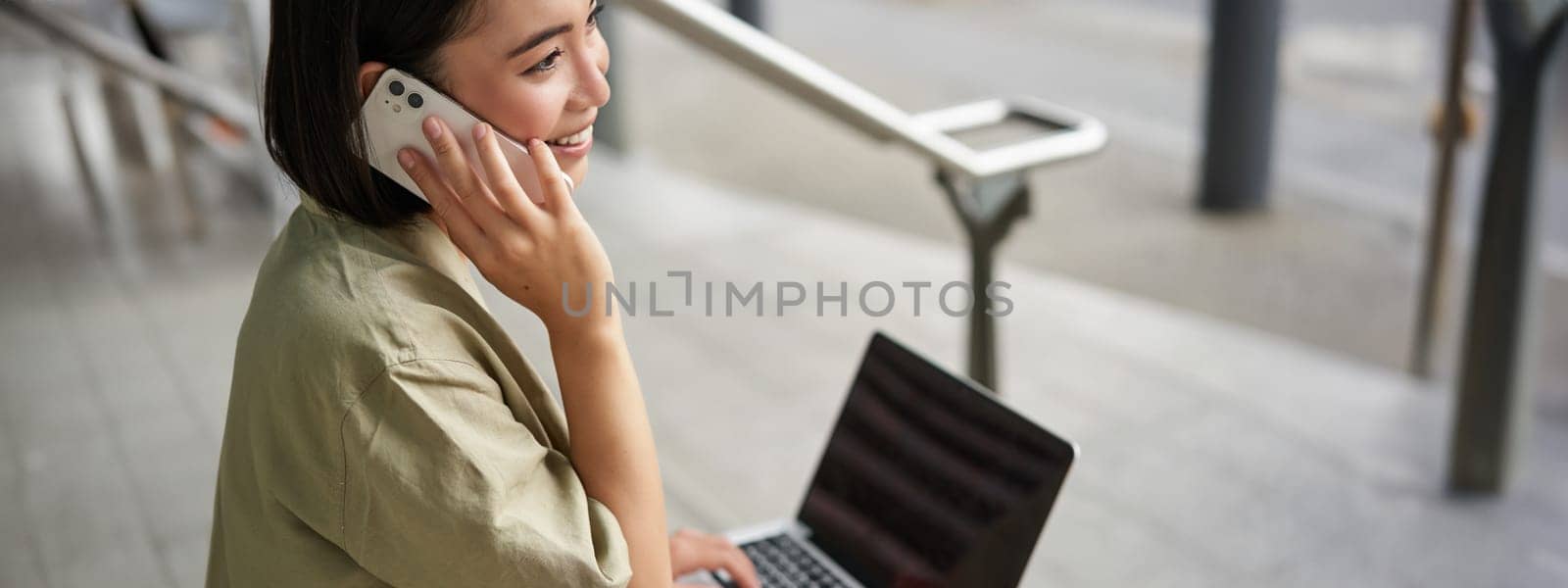 Portrait of asian girl, student sits on stairs with laptop, talks on mobile phone. Young woman makes a telephone call while working on computer.