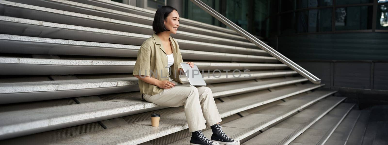 Young smiling asian girl with laptop, sits on street of city with coffee. Young woman does homework on computer while sitting on street stairs, digital nomad works remote.