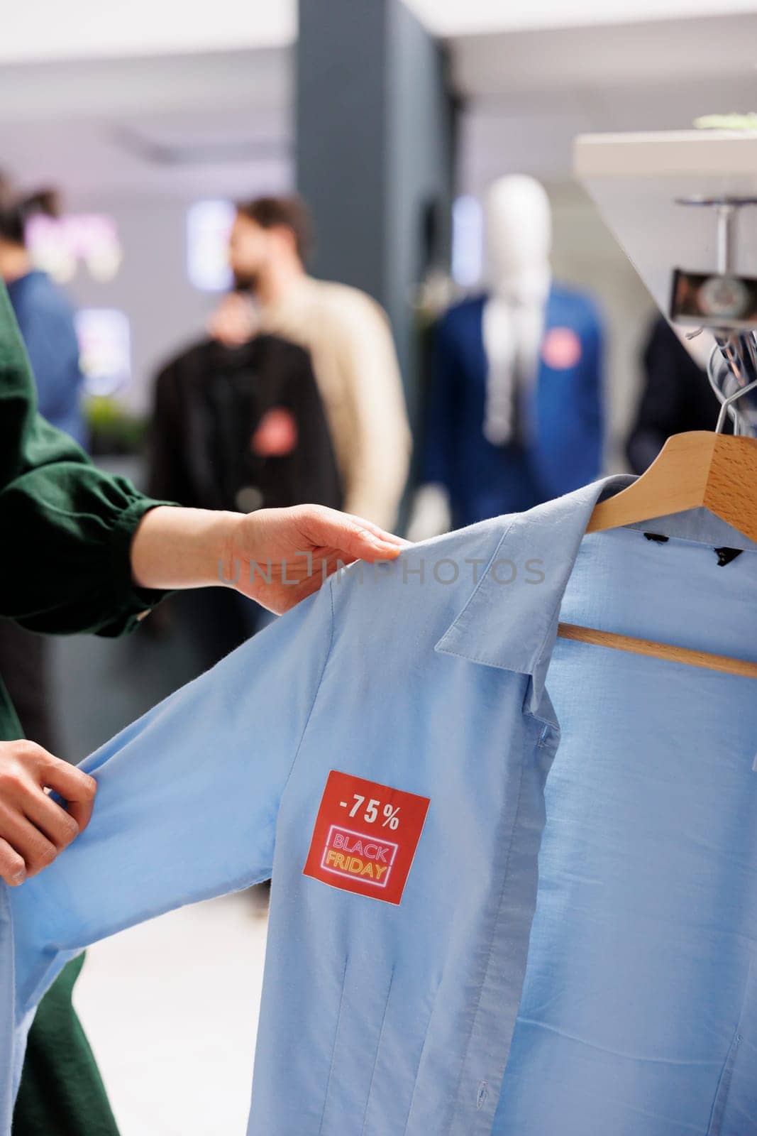 Woman customer looking through clothes hanging on rack, checking Black Friday offer in trendy Fashion boutique. Male shopper holding blue shirt with 75 percent discount red sale price tag