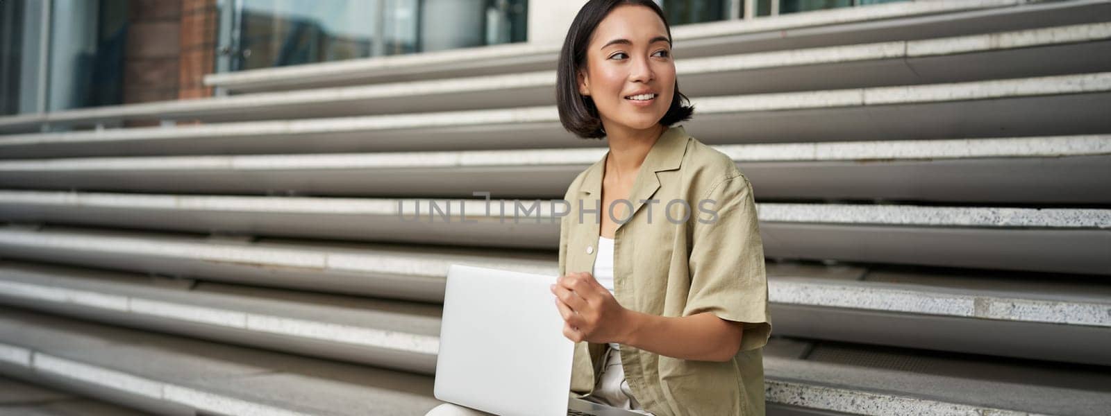 Portrait of young woman student, asian girl using laptop. Asign smiling girl, digital nomad works on her project remotely, sitting on street.