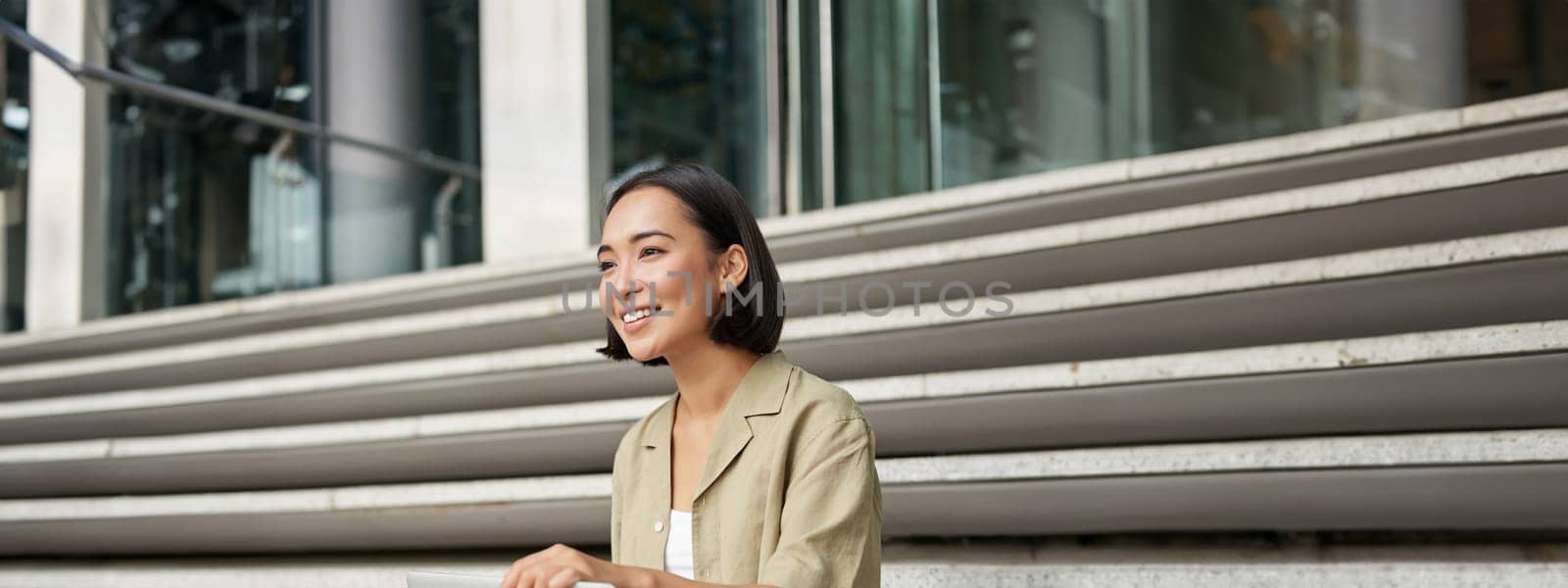 Vertical shot of smiling asian woman with laptop computer, sitting on stairs outside building. by Benzoix