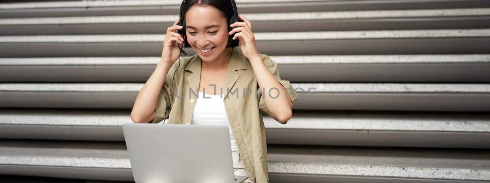 Portrait of asian woman, digital nomad girl using laptop and listens to music outdoors. Young student works on computer and smiles by Benzoix
