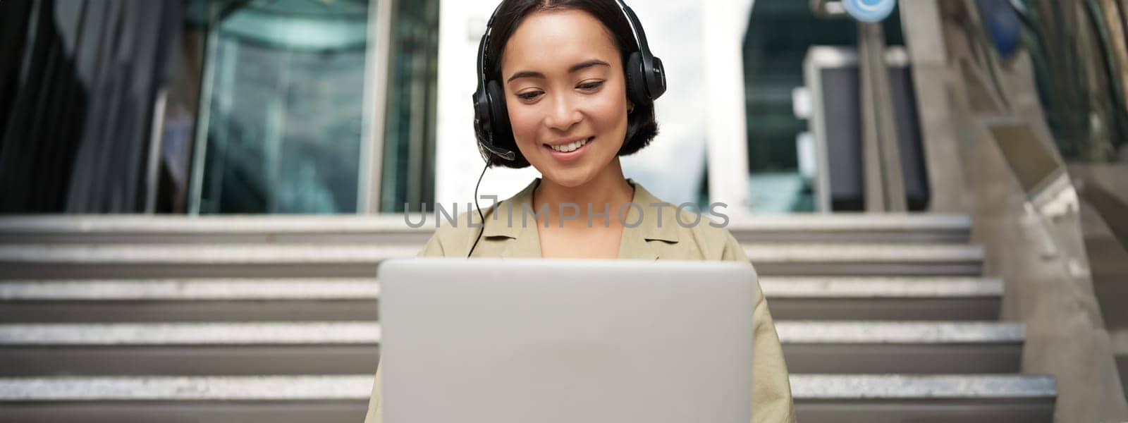 Portrait of young asian woman sitting with laptop and headphones, watching video, does online course on computer, sitting on stairs outdoors.