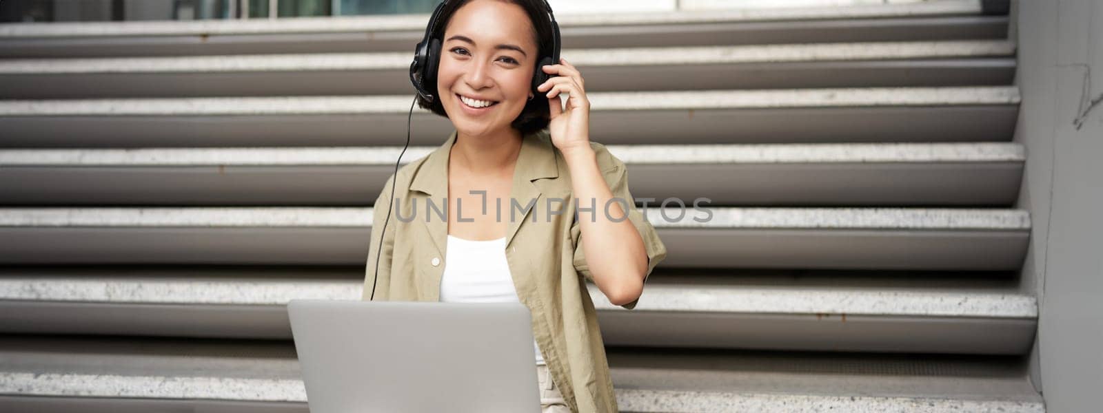 Smiling asian girl in headphones, works on laptop, digital nomad using computer on remote, sitting on stairs outdoors.