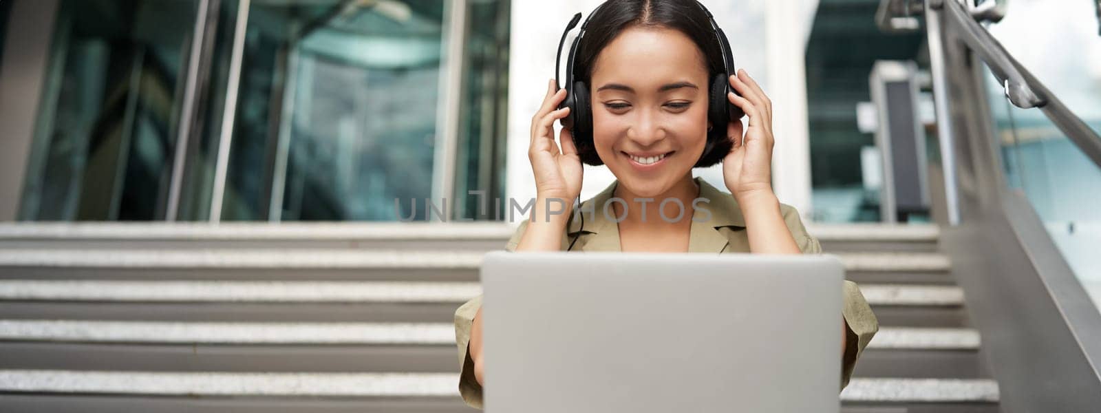 Portrait of young asian woman sitting with laptop and headphones, watching video, does online course on computer, sitting on stairs outdoors.
