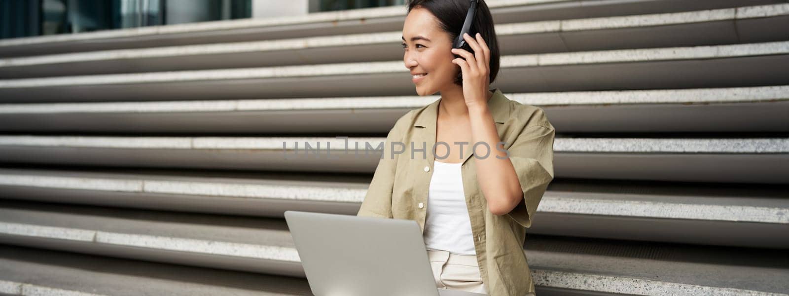 Portrait of stylish young woman smiles, sits on stairs with headphones and laptop, works on homework for university.