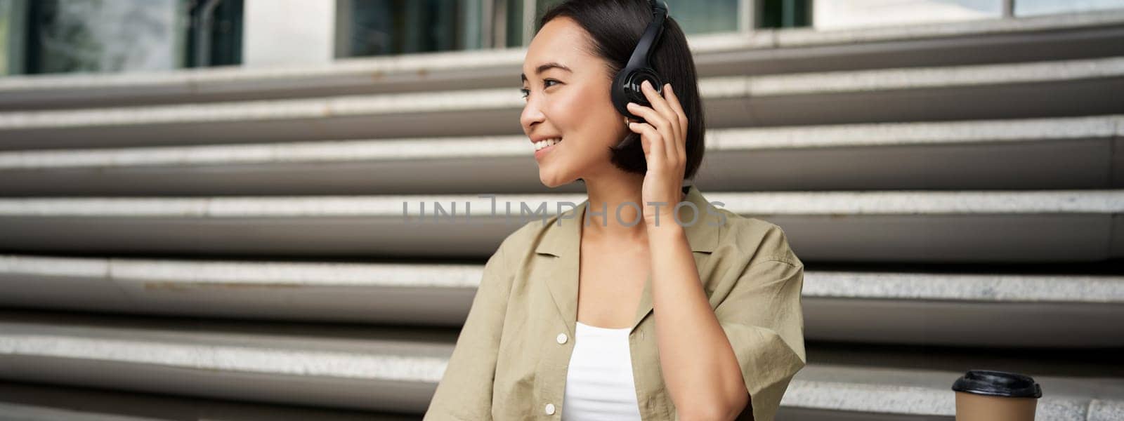 Close up portrait of smiling asian woman listens music in headphones, turns around with happy face expression, resting outdoors.