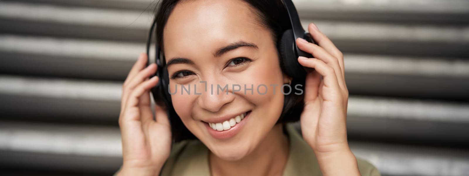 Close up portrait of smiling asian girl in headphones, listens to music outdoors, looking happy. People concept